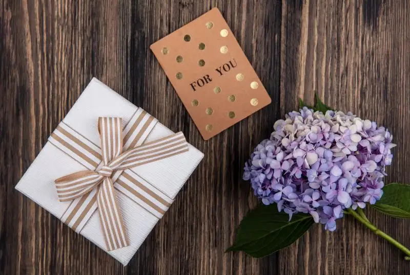 White gift box with striped ribbon, a 'For You' card, and a purple hydrangea on a wooden surface